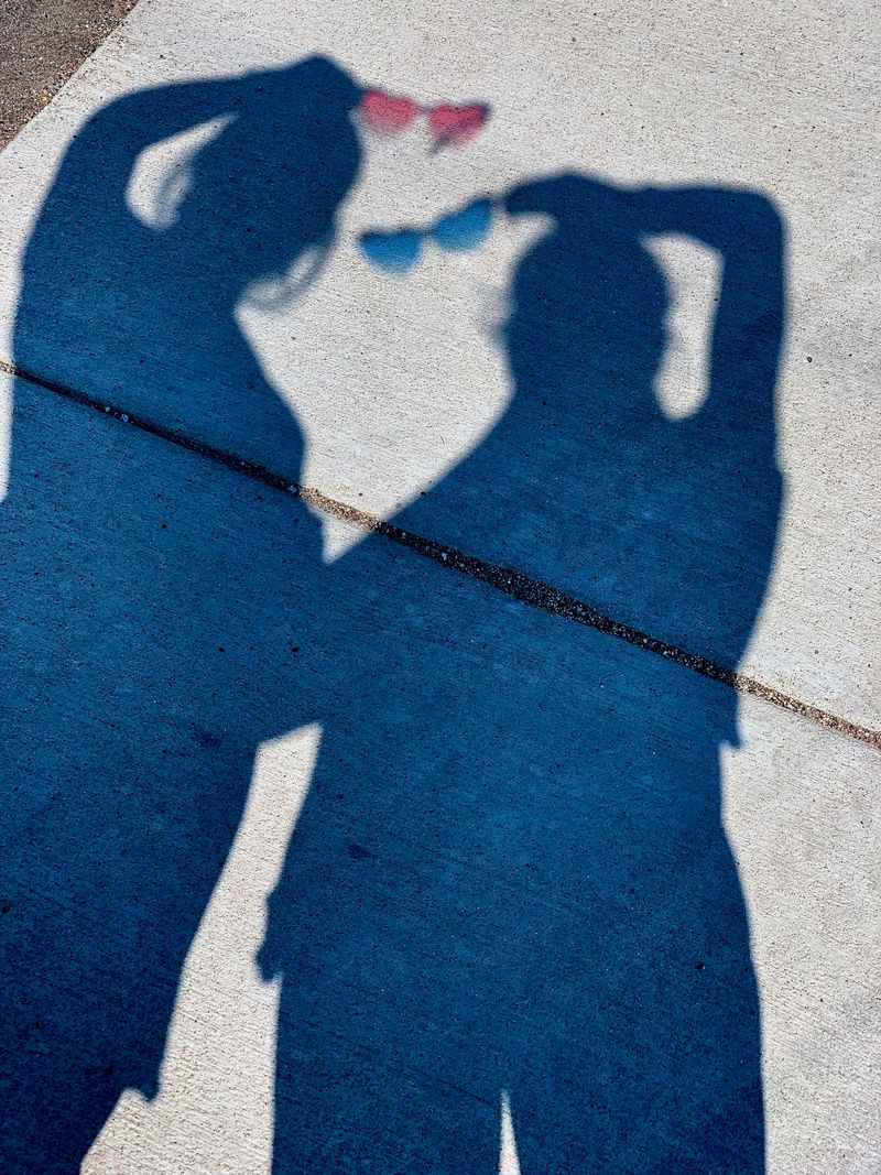 The sidewalk shadows of two people holding heart-shaped sunglasses up to sunlight.