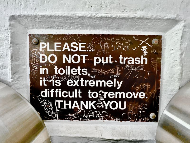 A sign that reads 'PLEASE DO NOT PUT TRASH IN TOILETS, IT IS EXTREMELY DIFFICULT TO GET OUT'.
